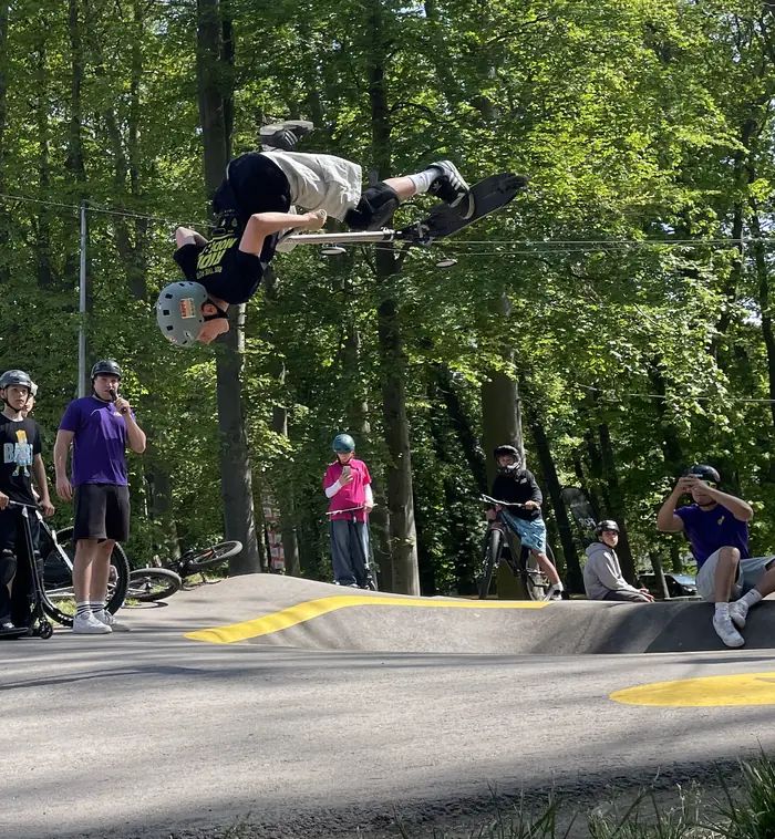A scooter rider performs a flip above the Darłowo pumptrack while participants and a presenter watch.